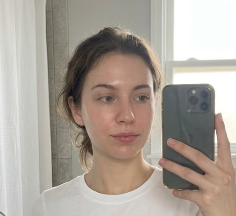 A woman taking a mirror selfie in a bathroom, wearing a white crewneck t-shirt and holding a dark gray smartphone.